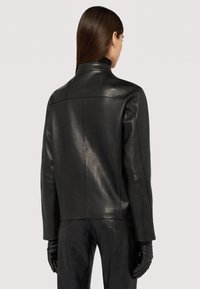 Schwarze Lederjacke mit hohem Kragen, glatter Textur und minimaler Naht. Kombiniert mit schwarzen Lederhandschuhen und -hosen.