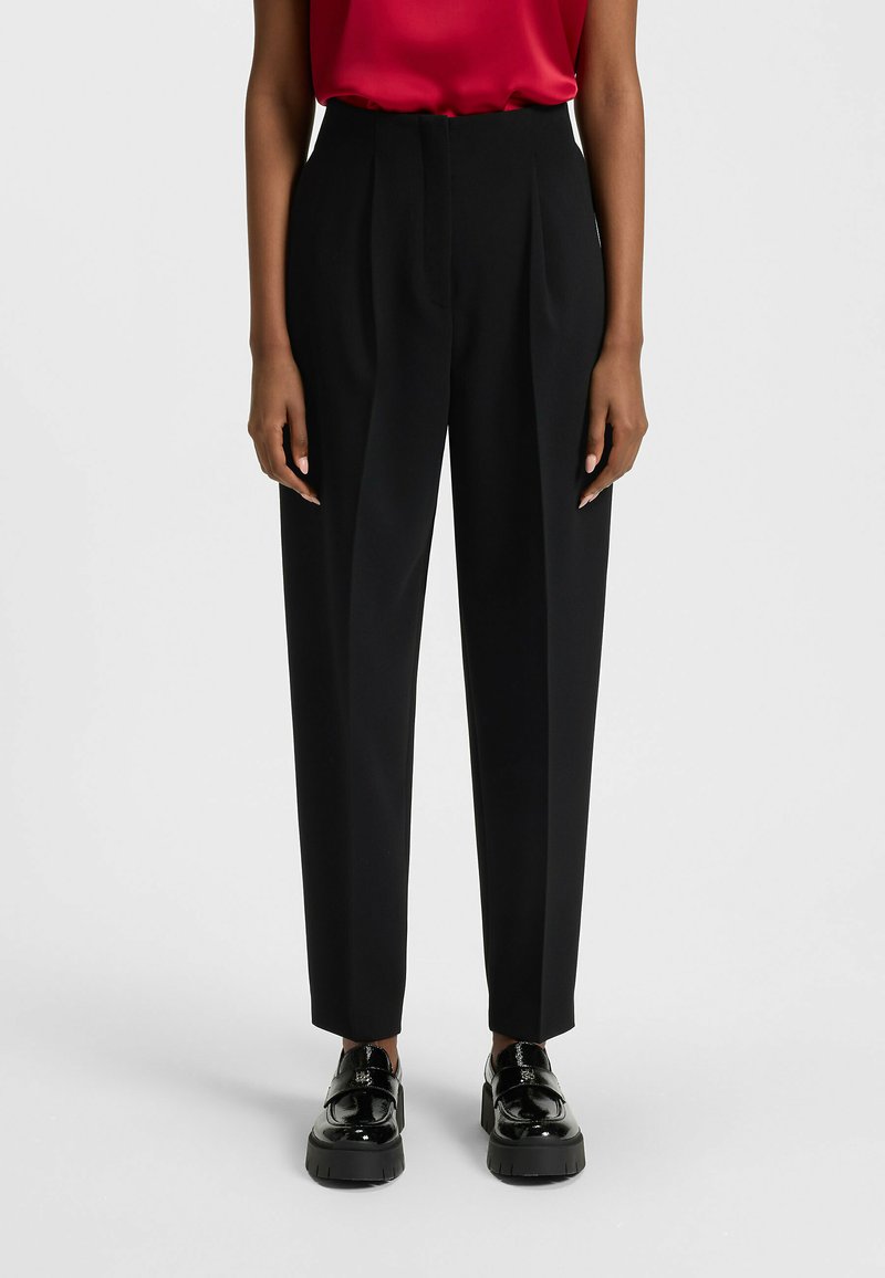 Black high-waisted trousers with a straight-leg cut, featuring front pleats, and paired with glossy black loafers. Smooth fabric texture.