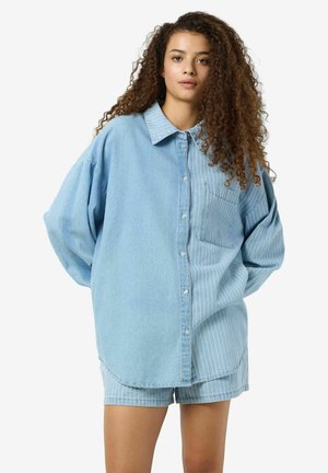 Camicia oversize in denimazzurro chiaro con design a righe, chiusura a bottoni e taschino singolo sul petto; abbinata a pantaloni corti a righe coordinati.