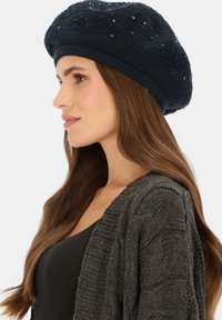 Granatowy dziergany beret z cekinowymi akcentami, okrągły kształt, teksturowane wykończenie oraz szeroki ściągacz, noszony pod kątem przez modelkę.