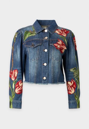 Personne portant une veste en jean bleue avec broderie florale rouge et blanche, un jean bleu, des bottes noires à talons, debout devant un fond uni.