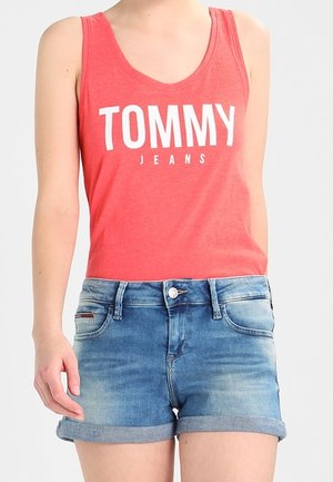 Kobieta w czerwonym topie bez rękawów z tekstem "TOMMY JEANS" i niebieskich podwiniętych dżinsowych szortach na jednolitym jasnym tle.