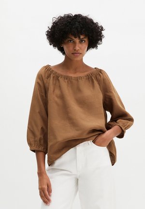 Donna con capelli ricci che indossa una blusa marrone ampia con le spalle scoperte e pantaloni bianchi, in piedi con una mano in tasca.