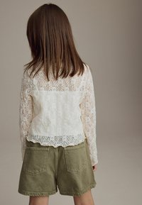 Hvid blonde langærmet top med bølget kant, parret med olivengrønne shorts med to baglommer, set fra ryggen.