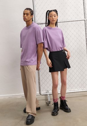 Tricou oversized lila, pantaloni largi bej, fustă neagră înfoiată cu curea, cizme negre cu șireturi și șosete roz cu un model gradient.