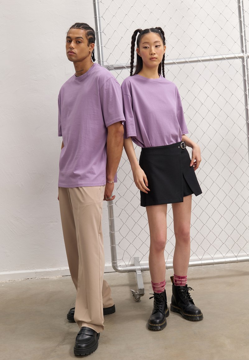 Tricou oversized lila, pantaloni largi bej, fustă neagră înfoiată cu curea, cizme negre cu șireturi și șosete roz cu un model gradient.