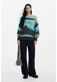 Maglione oversize lavorato a maglia con motivi astratti in teal, blu e beige, abbinato a pantaloni larghi navy e una borsa a spalla grigia.