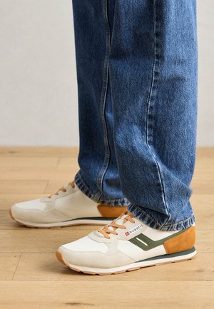 Białe i beżowe sneakersy z zielonymi akcentami, teksturowana tkanina i beżowe sznurówki. W zestawieniu z niebieskimi dżinsami na drewnianej podłodze.