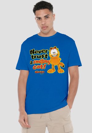 Blue cotton t-shirt featuring a cartoon cat and bold text saying "Never trust a smiling cat!" with a graphic logo at the bottom.