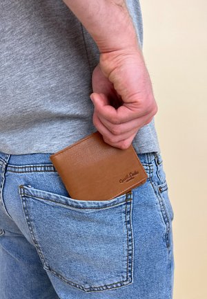 Brun læderpung delvist stoppet i en blå denimlomme, med synlig syning og præget mærkedetalje. Grå skjorte i baggrunden.