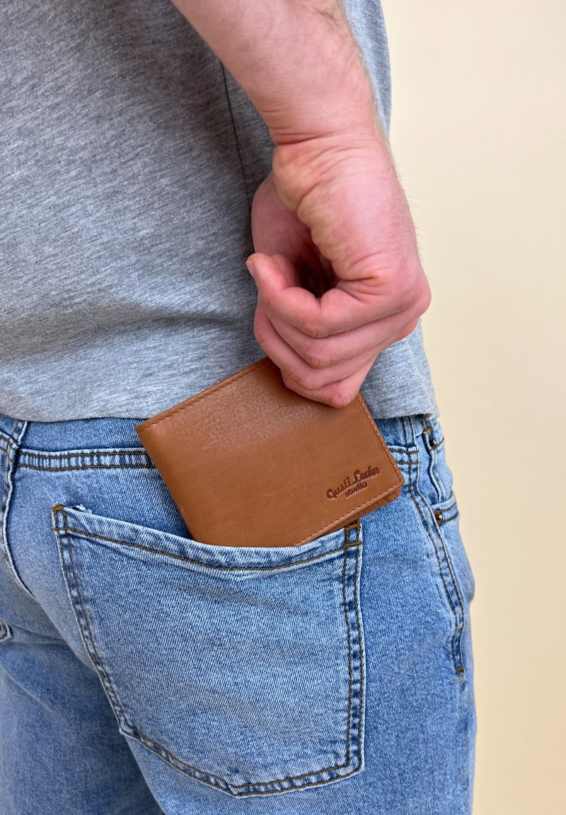Brun læderpung delvist stoppet i en blå denimlomme, med synlig syning og præget mærkedetalje. Grå skjorte i baggrunden.