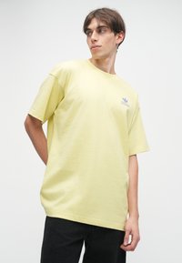 Κίτρινη βαμβακερή μπλούζα με άνετη εφαρμογή, στρογγυλή λαιμόκοψη και κοντά μανίκια. Διαθέτει μικρό μοβ λογότυπο της Adidas στο στήθος.