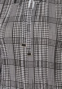 Tkanina w czarno-biały wzór houndstooth o gładkiej fakturze; na przodzie znajdują się dwa małe złote akcenty jako detal.