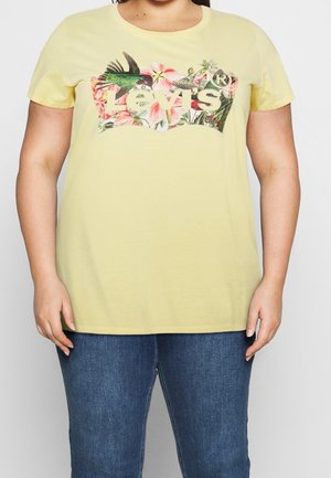 Personne portant un t-shirt jaune Levi's avec un logo floral et colibri, assorti à un jean bleu, debout devant un fond uni.