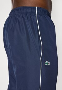 Pantaloni sportivi navy con vita elastica, piping bianco laterale e logo di un coccodrillo verde ricamato sull'orlo sinistro. Tessuto leggero.