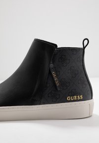 Zwarte enkelboot met een glad leren voorpaneel en een textiel achterpaneel. Bevat een gouden "GUESS" logo en een trekband. Beige rubberen zool.