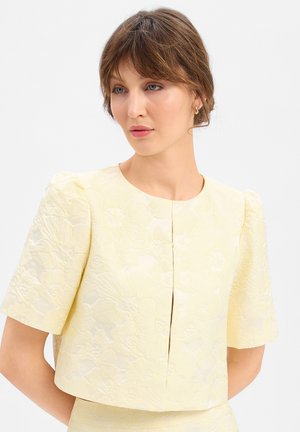 Vrouw met lichtbruin haar draagt een lichtgele, gebloemde, textuur korte mouw blouse en kijkt iets opzij tegen een witte achtergrond.