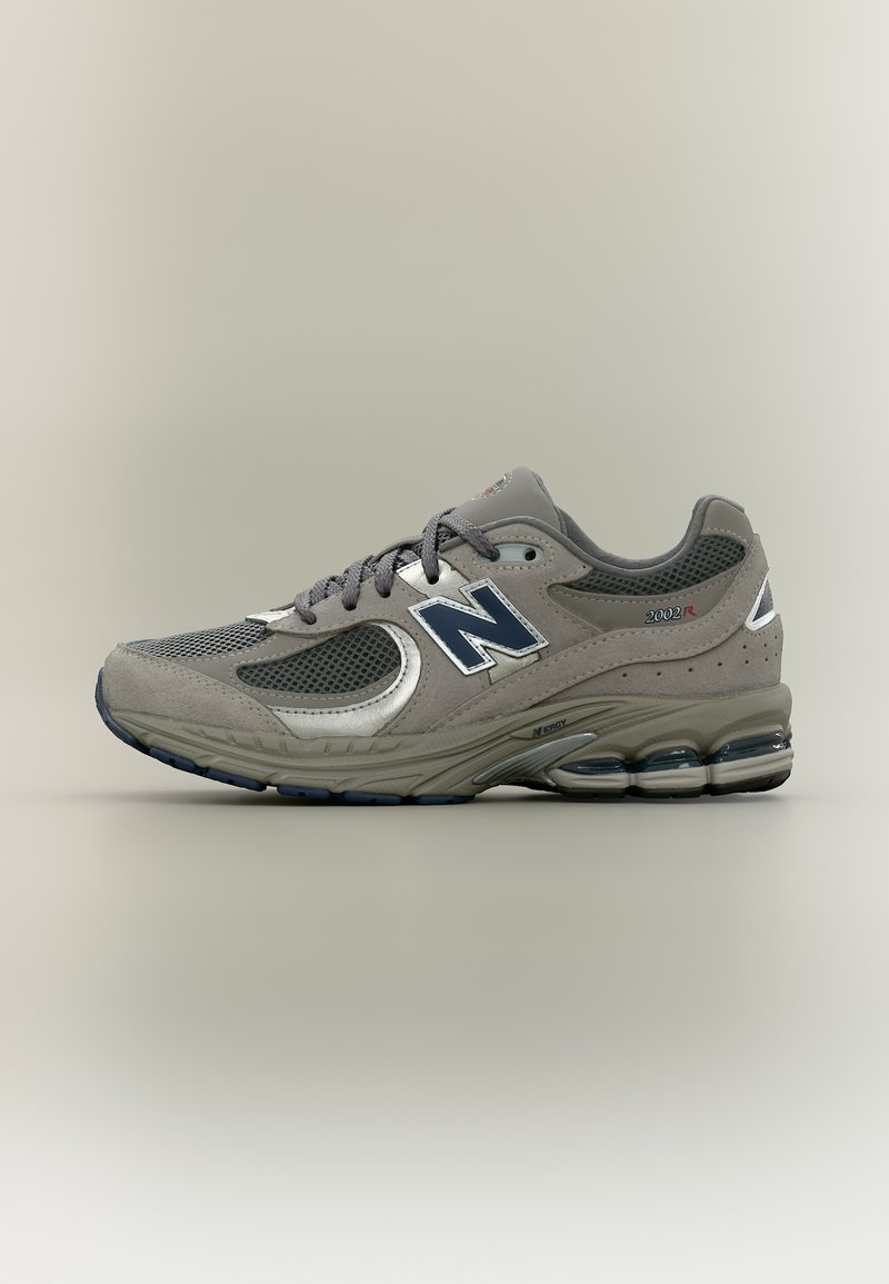 Γκρι παπούτσι New Balance 2002R με σκούρο μπλε λογότυπο, επάνω μέρος από διχτυωτό ύφασμα και σουέντ, μαλακή σόλα και ορατή αερόσολα στη φτέρνα.