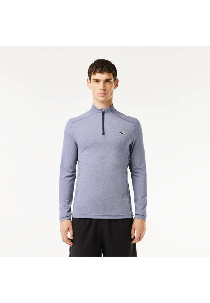 Hellblauer Pullover mit langen Ärmeln und Reißverschluss am Hals, mit einer strukturierten Oberfläche und einem dezenten Logo auf der Brust, kombiniert mit schwarzen Shorts.