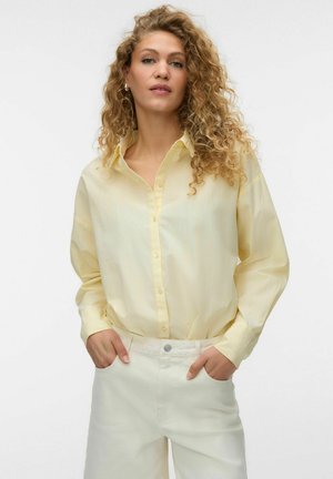 Kvinde med krøllet blondt hår iført en lys gul skjorte med knapper og hvide bukser, hænderne i lommerne, stående mod en ensfarvet baggrund.