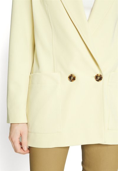 Κίτρινο διπλόθωρο σακάκι φτιαγμένο από υφή ύφασμα, διαθέτοντας δύο μεγάλες τσέπες και κουμπιά από ταρταρούγα.