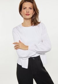 Weißer gerippter Cardigan mit lässiger Passform, langen Ärmeln und offener Vorderseite, kombiniert mit einem weißen Oberteil und schwarzen maßgeschneiderten Hosen.