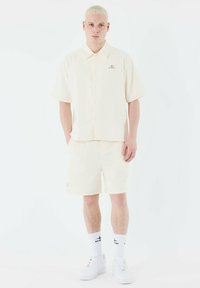 Camisa blanca de manga corta con relieve y pantalones cortos a juego. Cuenta con un cierre de botones, un logo sutil en el pecho y un corte holgado. Zapatillas blancas.