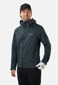 Homme portant une veste zippée bleu foncé avec capuche, casquette noire, pantalon noir et un gant blanc à la main droite, regardant légèrement vers la gauche.