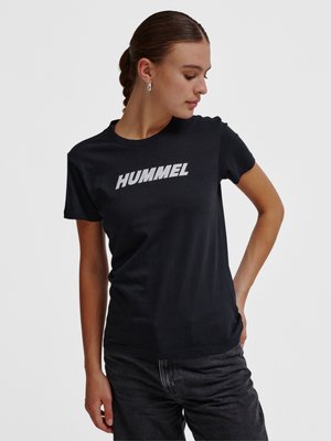 Jeune femme aux cheveux tressés portant des boucles d'oreilles argentées en forme de créoles, un t-shirt noir avec le logo "HUMMEL" et un jean foncé, regardant vers le bas à sa gauche.
