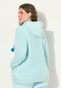 Chaqueta con capucha de color azul claro, fabricada en un tejido suave y texturizado. Presenta un detalle de pompón, mangas largas y dos bolsillos laterales.