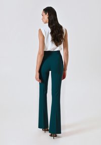Vrouw met lang golvend donker haar, in een mouwloos wit topje, teal hoge taille flare broek en zwarte hoge hakken, staande tegen een effen achtergrond.