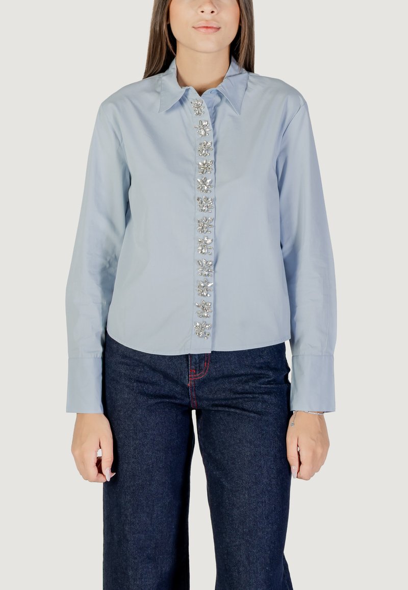 Camicia azzurro chiaro con colletto e bottoniera, decorata con elementi a forma di fiore, e maniche lunghe, abbinata a pantaloni in denim scuro.