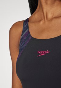 Maillot de bain noir avec un motif diagonal violet et bleu sur l'épaule, avec un logo Speedo rose sur la poitrine. Tissu lisse et extensible.