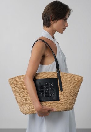 Femme en robe blanche sans manches portant un grand sac cabas tissé beige avec un patch en cuir noir et une lanière sur l'épaule.