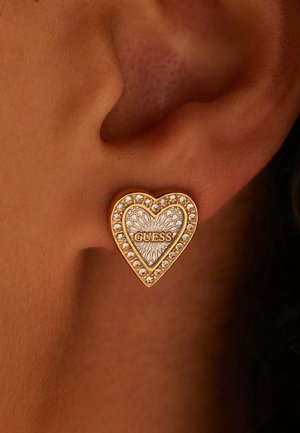 Gouden hartvormige oorbellen met een gestructureerd oppervlak, versierd met heldere rhinestones, met het "GUESS"-logo in het midden.