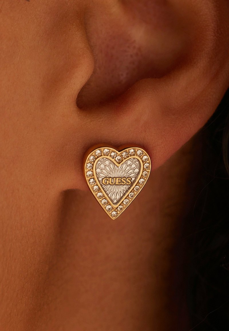 Orecchini a forma di cuore in tono oro con superficie texturizzata, impreziositi da strass trasparenti, con il logo "GUESS" al centro.