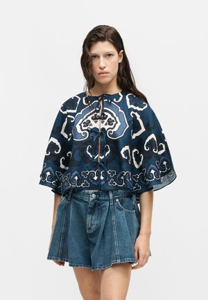 Femme portant un chemisier bleu marine à motifs avec des liens à l'avant, associé à un short en jean plissé taille haute, sur un fond clair uni.