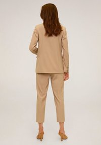 Femme aux cheveux auburn portant un tailleur beige avec un pantalon court et des mules à talons hauts, se tenant devant un fond clair uni.