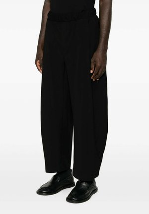 Uomo con pantaloni larghi neri e scarpe nere in pelle senza lacci, in piedi su uno sfondo bianco semplice.