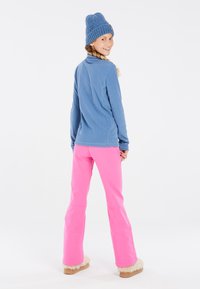 Maglione in pile blu con colletto alto, abbinato a pantaloni da sci rosa brillante con gambe svasate e orlo foderato in pelliccia sintetica. Berretto lavorato a maglia blu.