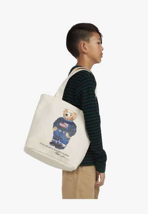 Cremefarbene Canvas-Tasche mit einem grafischen Druck eines Teddybären, der einen Pullover mit amerikanischer Flagge und Jeans trägt. Verfügt über zwei Schultertragegurte.
