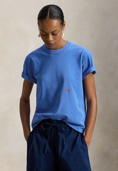 Γαλάζια βαμβακερή μπλούζα με ριγμένα μανίκια, στρογγυλή λαιμόκοψη και κόκκινη κέντημα "Polo" στο στήθος, συνδυασμένη με ναυτικά παντελόνια με κορδόνι στη μέση.