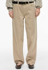 Pantalon beige à jambes larges en tissu au toucher lisse. Dispose d'une ceinture classique et de deux poches avant. Porté avec une ceinture noire.