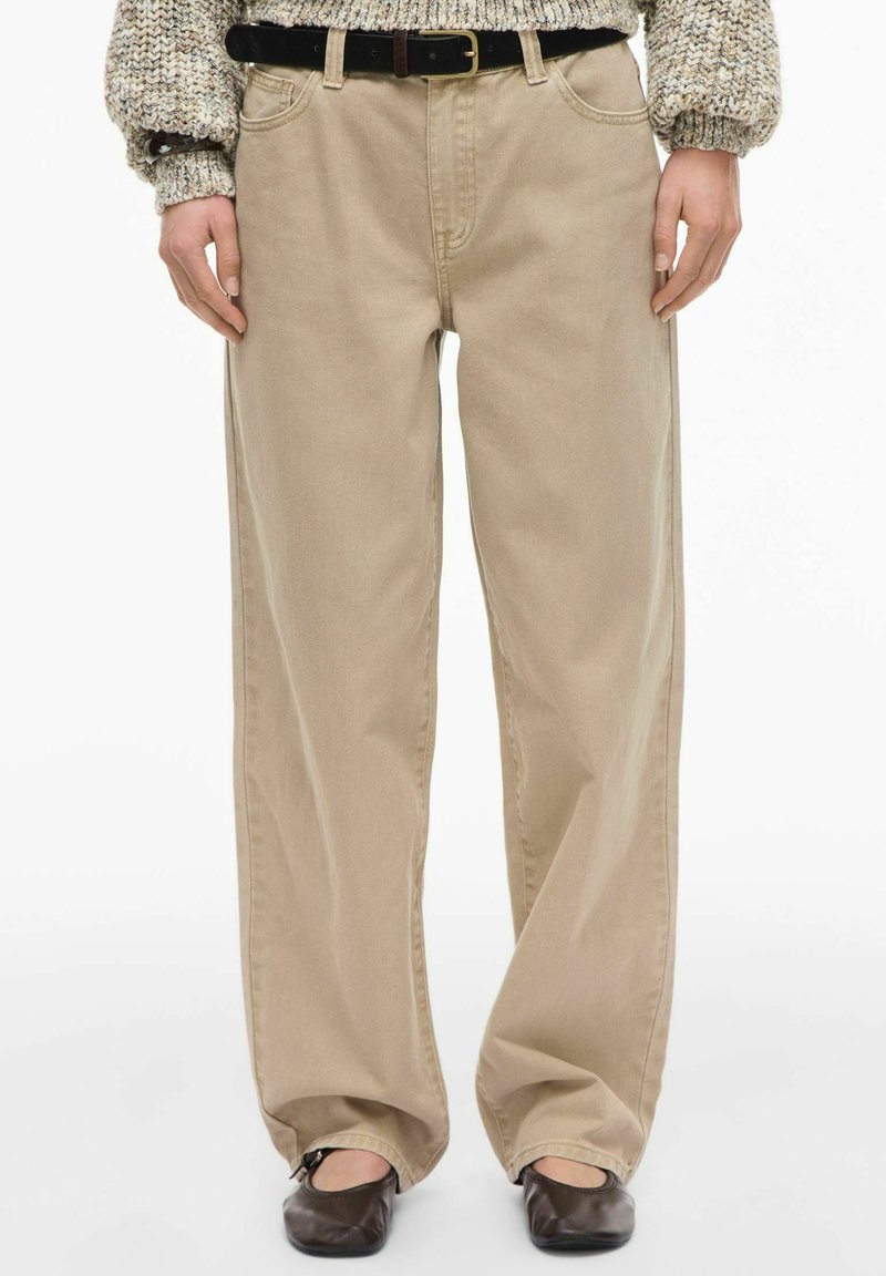 Pantalon beige à jambes larges en tissu au toucher lisse. Dispose d'une ceinture classique et de deux poches avant. Porté avec une ceinture noire.