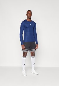 Kék, hosszú ujjú sportfelső Adidas logóval; szürke nadrág fekete és szürke csíkokkal; fehér térdig érő zoknik; fehér sportcipő.