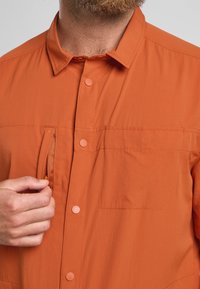 Oranje shirt met een button-down kraag, vóórsluiting en borstzak. Heeft een gestructureerde stof met een ritszakdetail.