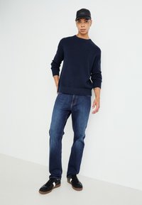 Jūras spēku adīts džemperis ar ribotām piedurknēm, apvienots ar ziliem džinsiem un melniem sporta apaviem. Modelis valkā melnu cepuri, stāvot pret vieglu fonu.