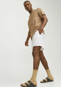 Chemise à manches courtes à motifs bruns, shorts blancs, chaussettes côtelées beiges, et sandales noires avec boucles, se tenant devant un fond neutre.