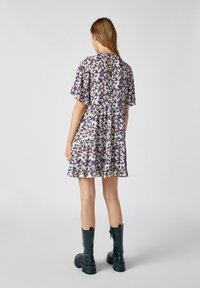 Robe courte florale avec manches volantées, ourlet à volants et dos en forme de goutte. Tissu aux tons crème, violet et bleu ; associée à des bottes noires montantes jusqu'au genou.