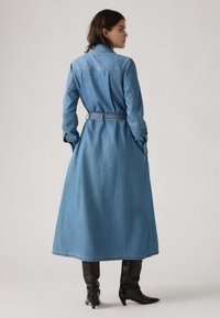 Fénykék denim trench coat övvel a deréknál, hosszú ujjal és egyenes szabással, fekete térd fölé érő csizmával párosítva.